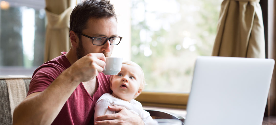 Hombre sentado y sujetando a una bebé con un brazo mientras toma café y trabaja