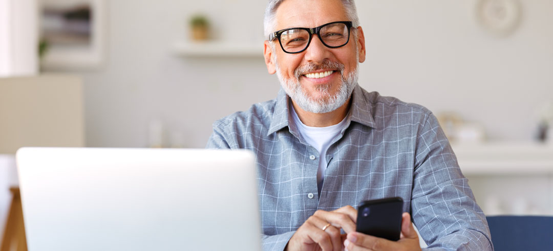 Hombre mayor sonriendo y consultando su teléfono móvil y el ordenador